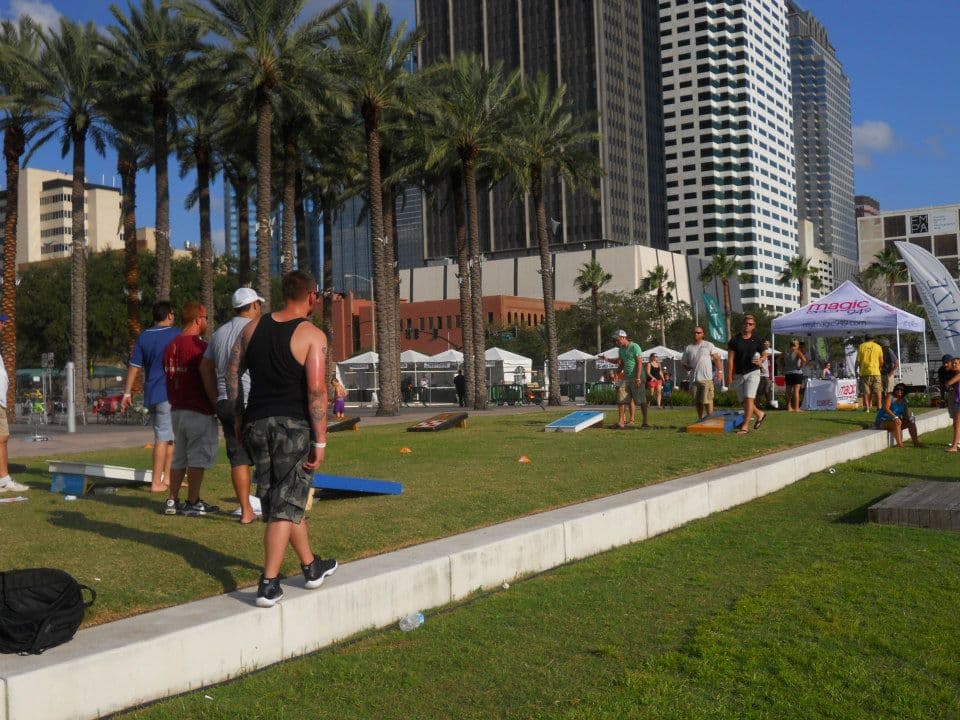 Players at an outdoor cornhole tournament with rows of regulation boards set up on a park lawn